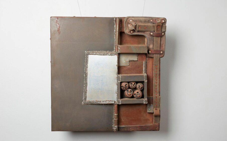 Square wall-mounted sculpture composed of mainly square and rectangle metal-looking shapes layered and soldered, creating a patchwork effect. The left half is dark grey mottled with rust color at the top edge. A vertical flat silver rectangle sits roughly in the center of the piece surrounded by jagged soldering. The right side consists of a rust-colored metal base topped with three-dimensional shapes that appear to be composed of leather strapping, more soldered metal shapes and spheres carved with faces. Dusty brown leather strapping punctuated with rivets creates an L shape starting at the top right corner running down the right edge. More riveted leather strapping continues on the bottom right edge. A metal box containing five spheres with faces with varying expressions sits two-thirds from the top of the sculpture touching the center line that divides the piece. Facial expressions include two smiling, one grimace, and two with what appear to be O-shaped mouths. Rough soldering creates thick L shapes in rust and gray above the box of spheres.
