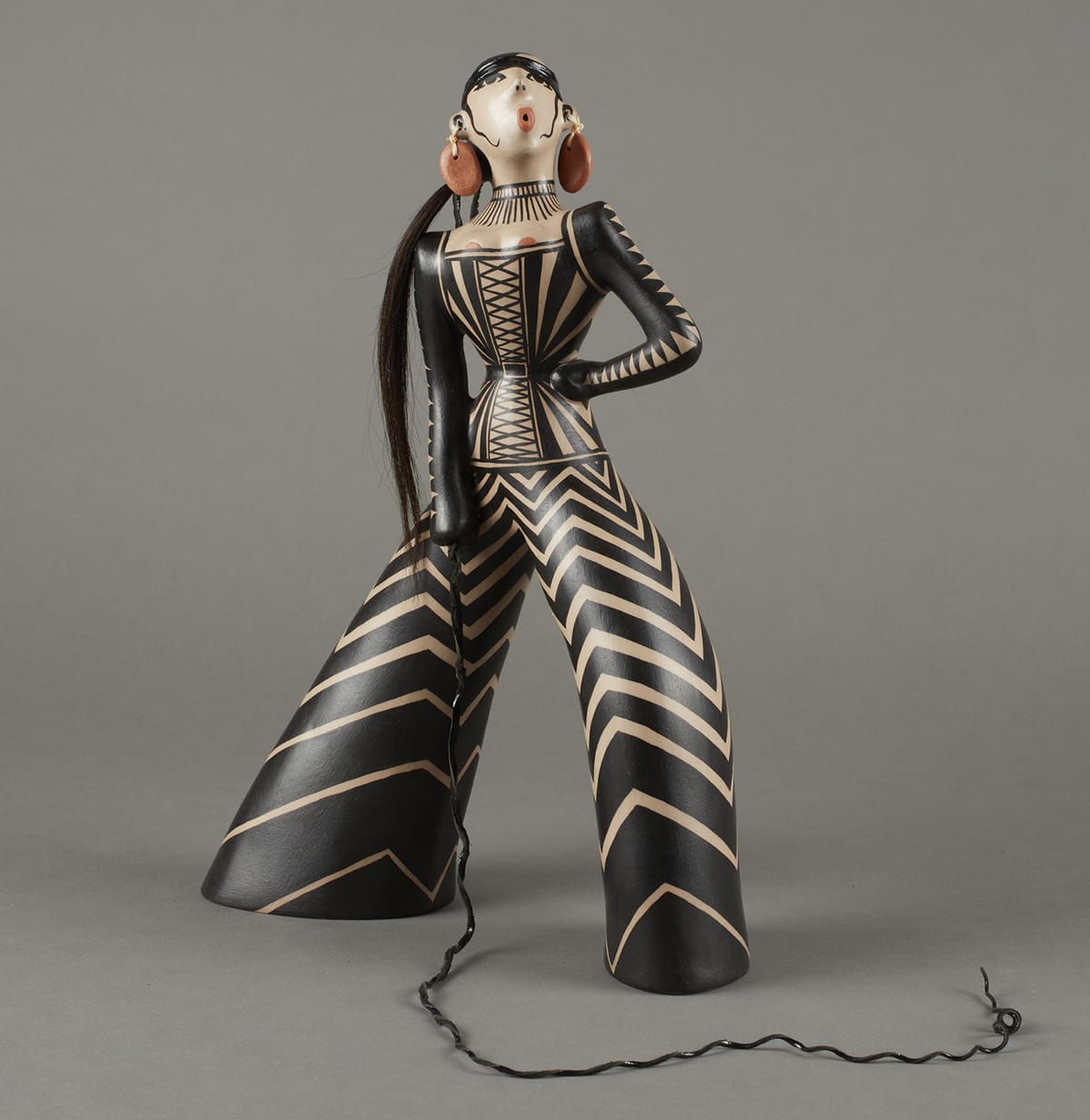 Stylized figure adorned with black-and-white triangular patterns. Details include reddish circular earrings similar to the shape of the lips, a long ponytail, and whip.