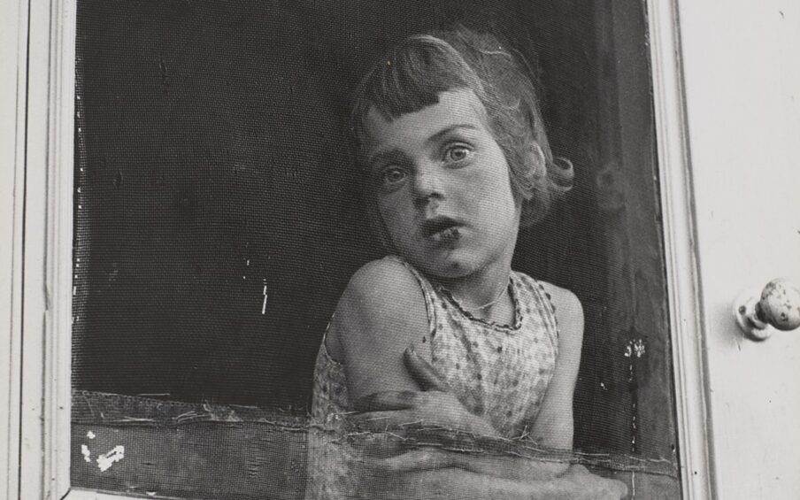 A vertical, rectangular, black-and-white photograph, featuring a young, light-skinned child at center. She leans, arms folded across her upper waist, on the inside of a patched and worn screen door, gazing through the screen and outside to the photographer/viewer. The interior space behind her is dark and without detail. The light gray door frame is seen along both the bottom and right edges of the image. A worn, chipped doorknob is on the door frame to the left of the figure at shoulder height. Her head is turned to the left, her chin touching her right shoulder. Her bobbed hair is chin-length with short, uneven bangs, and she wears a sleeveless, printed shirt. Dark areas appear on her mouth, nose and chin suggesting a scrape from a fall.