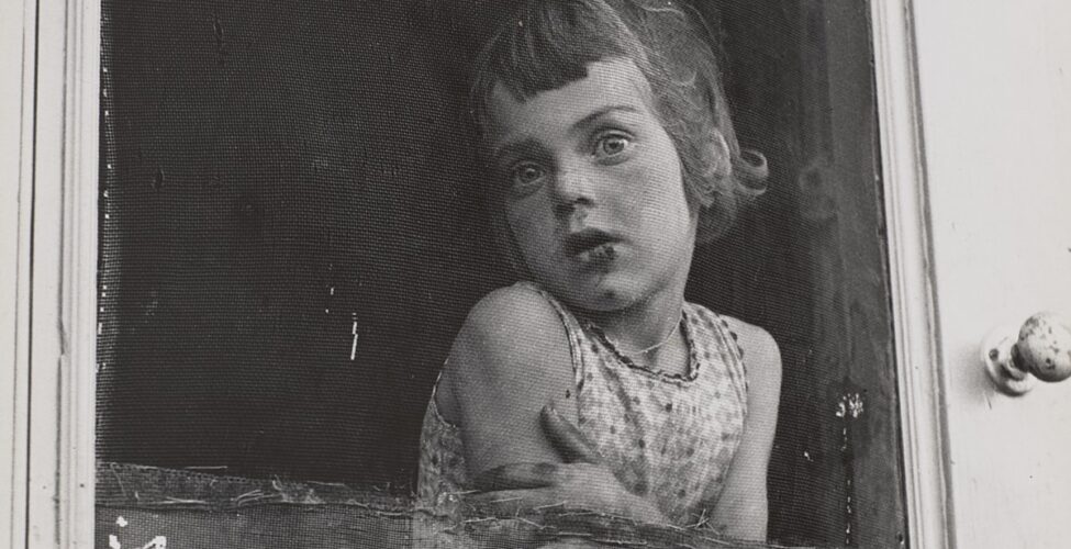 A vertical, rectangular, black-and-white photograph, featuring a young, light-skinned child at center. She leans, arms folded across her upper waist, on the inside of a patched and worn screen door, gazing through the screen and outside to the photographer/viewer. The interior space behind her is dark and without detail. The light gray door frame is seen along both the bottom and right edges of the image. A worn, chipped doorknob is on the door frame to the left of the figure at shoulder height. Her head is turned to the left, her chin touching her right shoulder. Her bobbed hair is chin-length with short, uneven bangs, and she wears a sleeveless, printed shirt. Dark areas appear on her mouth, nose and chin suggesting a scrape from a fall.