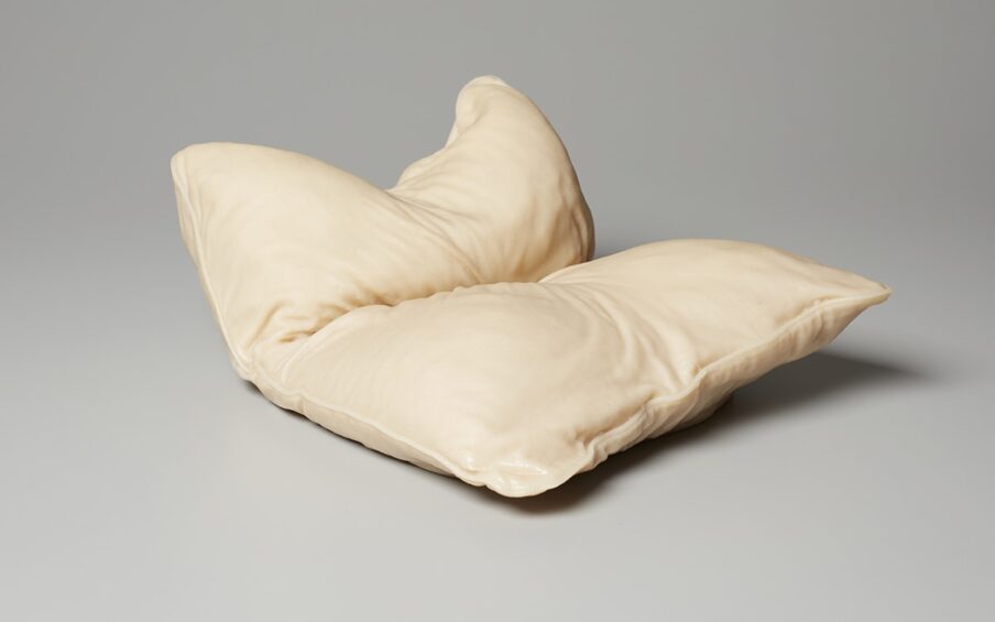 This sculpture depicts an object that resembles a bed pillow. Its shorter side is closest to the viewer. The sculpture is turned so one pillow corner is nearest the bottom center and the pillow is shown in a three-quarter view. It is deeply creased in the center as if it has been folded in half, short end to short end. The rear part of the pillow appears to be bunched upright and is indented in the center, corners jutting upward. Wrinkles, creases and piping along the seams are visible. The color is a very pale tan with cream highlights and deeper tones of tan in the creases. A shiny highlight of white is at the pillow corner nearest the viewer at bottom center.