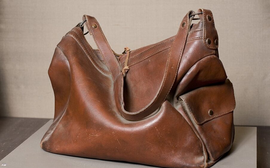 Photo of a sculpture representing a deep brown leather handbag. Viewed from the side, a rectangular handbag with both ends and middle collapse inward as if half empty. A highlight on the outward bulge of the main body of the bag is represented in a lighter color. A shoulder strap lies draped across the main body and lies next to the highlight. Hardware and rivets attach the strap to the main body of the bag at each end. A flat pocket with a flap is on the right end of the bag. Seams and stitching are clearly visible and a zipper puller is at the top center of the bag.