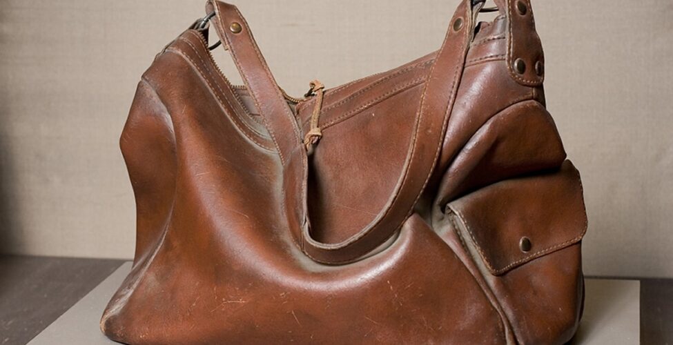 Photo of a sculpture representing a deep brown leather handbag. Viewed from the side, a rectangular handbag with both ends and middle collapse inward as if half empty. A highlight on the outward bulge of the main body of the bag is represented in a lighter color. A shoulder strap lies draped across the main body and lies next to the highlight. Hardware and rivets attach the strap to the main body of the bag at each end. A flat pocket with a flap is on the right end of the bag. Seams and stitching are clearly visible and a zipper puller is at the top center of the bag.