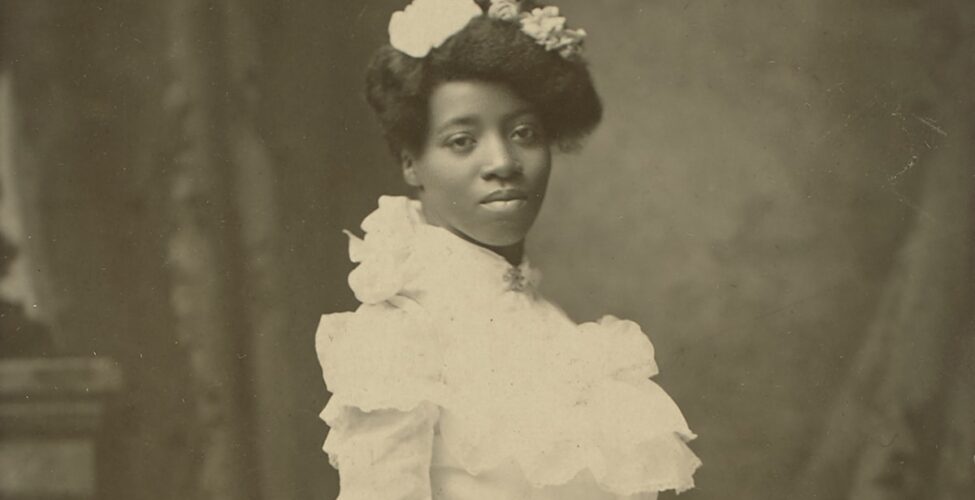 A black-and-white photograph of a young African American woman standing turned to the right in a three-quarter view, head facing forward, gazing directly at the viewer. She wears a white, floor-length gown with high neck and long sleeves. Her hair is swept up, piled on the crown of her head, and adorned with several flowers. Her expression is neutral, her head slightly tilted to the left. She holds a large, full bouquet of roses, carnations, and ferns whose long stems are tied with a ribbon. The flowers are angled down toward the floor while the stems point up and away from the viewer. The bottom of her gown ends in several tiers of ruffled flounces. Behind the figure is a painted backdrop depicting drapery and part of a stone mantel. A wide, tan border surrounds the long, rectangular portrait, showing bumped and creased corners and staining at bottom. A handwritten inscription reads: “1st blk. graduate // from high school // in Hastings, Nebraska // about 8 yrs. before grandma Nonie”. A logo reading “Payne Hastings, Nebr” is printed in the bottom right corner of the border.