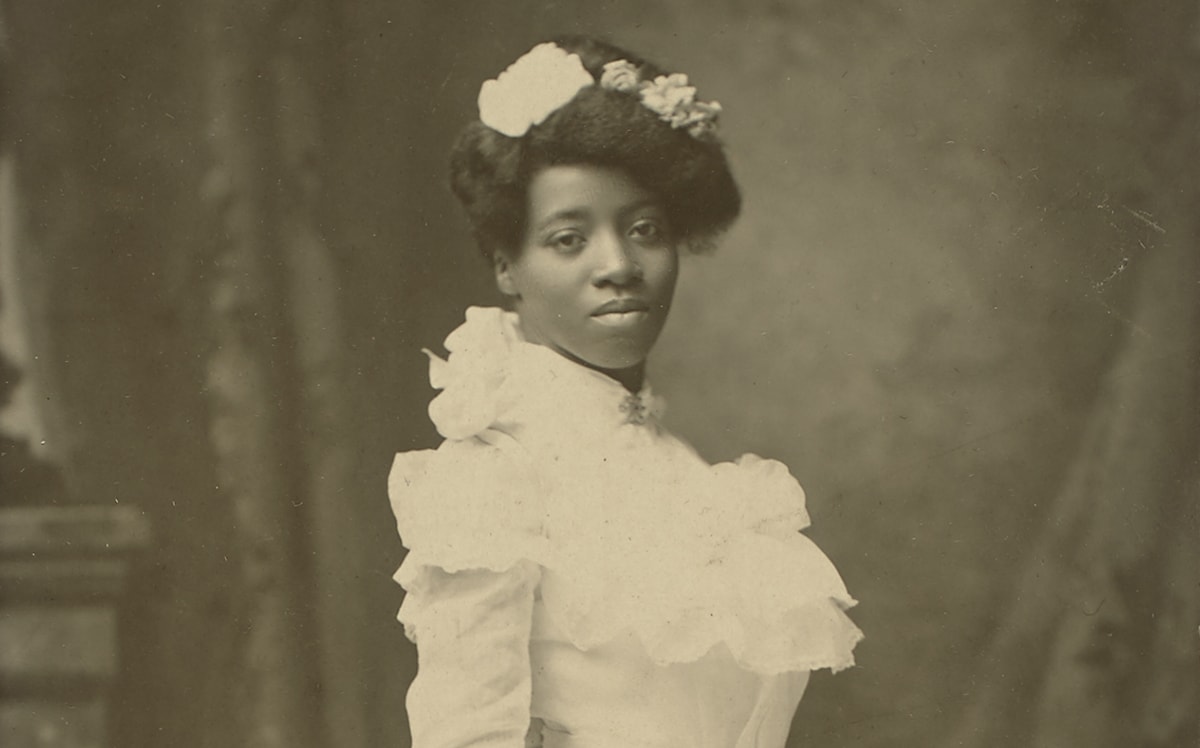 A black-and-white photograph of a young African American woman standing turned to the right in a three-quarter view, head facing forward, gazing directly at the viewer. She wears a white, floor-length gown with high neck and long sleeves. Her hair is swept up, piled on the crown of her head, and adorned with several flowers. Her expression is neutral, her head slightly tilted to the left. She holds a large, full bouquet of roses, carnations, and ferns whose long stems are tied with a ribbon. The flowers are angled down toward the floor while the stems point up and away from the viewer. The bottom of her gown ends in several tiers of ruffled flounces. Behind the figure is a painted backdrop depicting drapery and part of a stone mantel. A wide, tan border surrounds the long, rectangular portrait, showing bumped and creased corners and staining at bottom. A handwritten inscription reads: “1st blk. graduate // from high school // in Hastings, Nebraska // about 8 yrs. before grandma Nonie”. A logo reading “Payne Hastings, Nebr” is printed in the bottom right corner of the border.