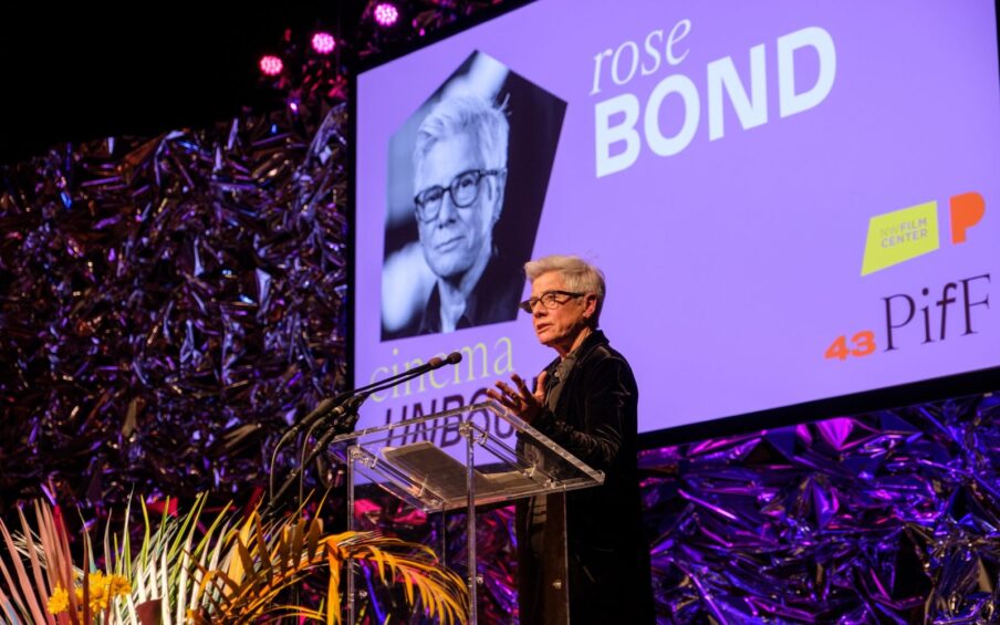 Photo of Rose Bond standing at a clear podium with a large screen in the background. Rose has short gray hair, wears black glasses, and is wearing a dark button up shirt and a large dark coat. Rose's left hand out outstretched toward the mic. A large fern grows in front of the podium. The purple screen behind has a photo of rose and reads rose BOND. cinema UNBOUND NW Film Center 43 PIfF.