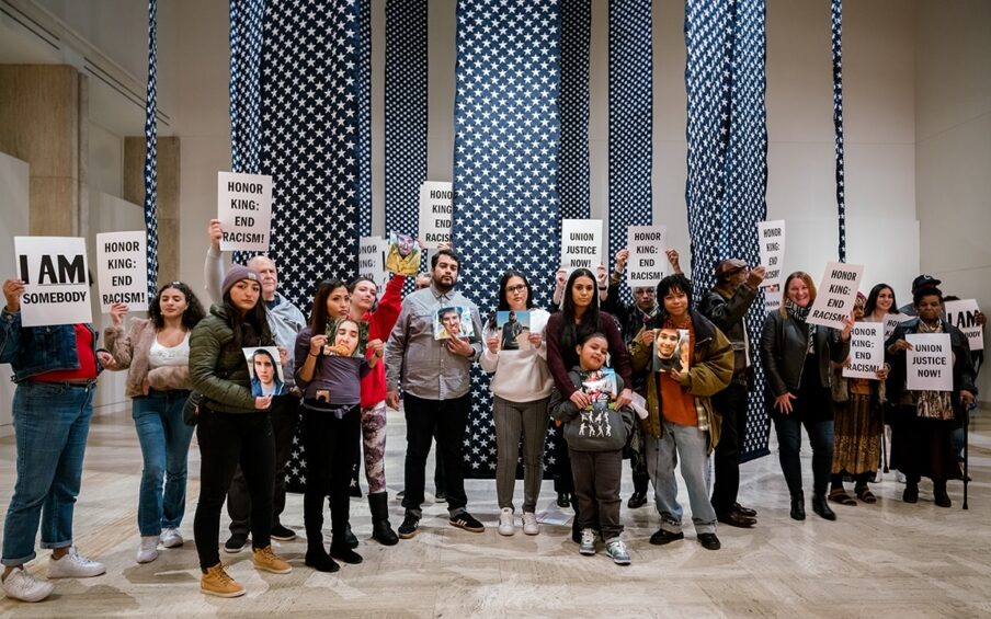 20 people of varying ages, races, and genders stand in museum gallery holding signs that read I AM Somebody, Honor King: End Racism, and Union Justice Now! They stand in front of a Hank Willis Thomas installation titled "14,719" which is a series of 28 foot tall blue banners hanging from the ceiling covered in white embroidered stars representing gun deaths in the US in 2018.