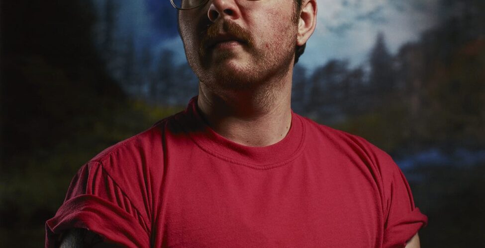 A vertical rectangular portrait of a masculine-presenting individual shown from the midsection up. The figure is positioned to face the viewer or artist and their head is turned to the left. They have light skin, dark short hair, a mustache, and beard stubble. They wear metal aviator-style eyeglasses and a red short-sleeved t-shirt with the sleeves rolled up. The figure’s bare arms show several tattoos. The upper arm at left has the words “MOM” and “DAD” within a heart design and another is of an insect. On the figure’s forearm at right, part of a bird tattoo is visible. The background is slightly out of focus and depicts large, jagged, snow-covered mountain peaks in blues and whites. Below the mountains are pine trees and dark foliage.