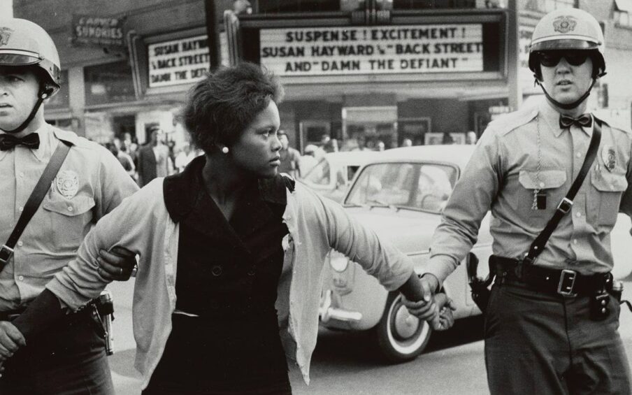 A horizontal rectangular black-and-white photograph of a young Black woman being detained by two male police officers. The woman is at center, flanked by the officers. Each holds the woman by her outstretched arms. The woman faces front but her head is turned to the right. She has dark skin, short wavy hair and wears a bright white, round earring. Her expression is neutral. Over her dark dress, she wears a light-colored cardigan sweater which is torn at the shoulder and front. The officers wear their police uniform of light shirts, dark pants, helmets, dark bow ties, duty belts with straps crossing the chest from shoulder to belt, holsters, and guns. The officer at left uses two hands to hold the woman’s arms, one hand on her upper arm and one hand on her wrist. The officer at right holds the woman’s wrist. Behind the three figures we see cars of the time, and a large movie marquee. The marquee reads “SUSPENSE! EXCITEMENT! SUSAN HAYWARD IN ‘BACK STREET ‘ AND ‘DAMN THE DEFIANT.' " People are crowded beneath the marquee.