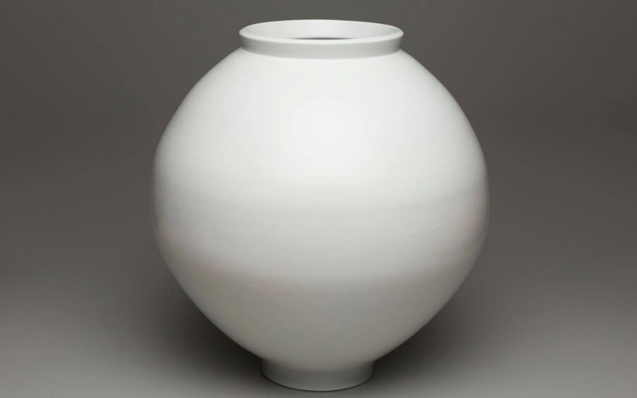 A photograph of soft-white, spherical-shaped jar that is subtly elongated vertically. It has a raised, upright lip that flares ever so slightly on a wide mouth. The base or foot is narrower than the mouth but mirrors its simple shape. Subtle striations run horizontally around the body of the jar giving the appearance of very faint texture. The jar is pictured against a gray background.