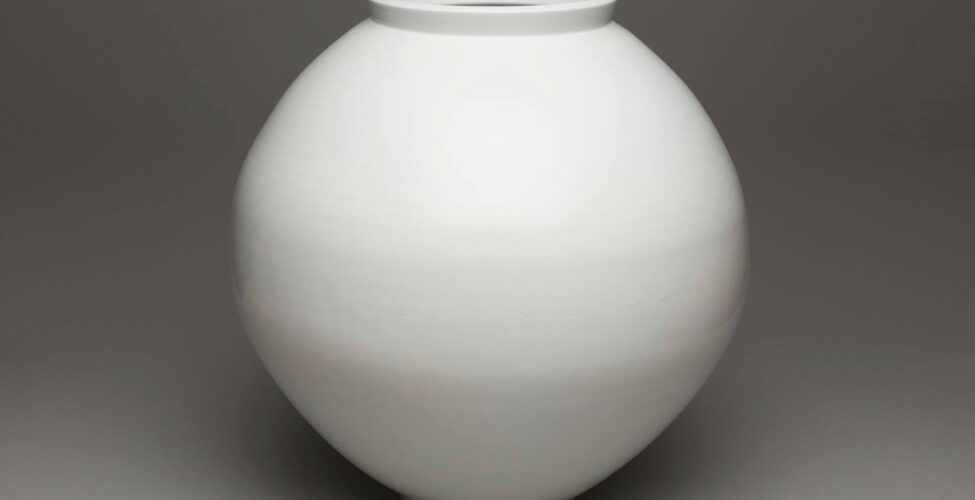 A photograph of soft-white, spherical-shaped jar that is subtly elongated vertically. It has a raised, upright lip that flares ever so slightly on a wide mouth. The base or foot is narrower than the mouth but mirrors its simple shape. Subtle striations run horizontally around the body of the jar giving the appearance of very faint texture. The jar is pictured against a gray background.