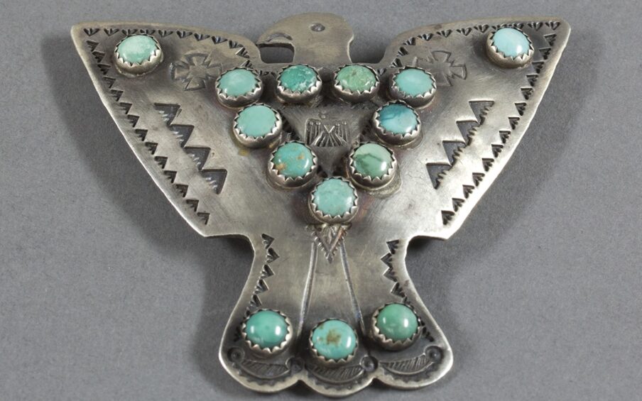 A photo of a silver bird shaped pin with wings open to each side. Its head and beak are shown in profile, facing left. The body of the bird is adorned with round turquoise stones set in an inverted triangle pattern. In the center is a small stamp of a thunderbird with its wings pointed down, head and beak facing right. Two small, stylized crosses are positioned on either side of the turquoise triangle. Just below this triangle is a small, stamped, inverted triangle with detailed decoration. There are single turquoise stones at the tip of each wing. The tail is depicted by three feathers with rounded tips and accented with three more turquoise stones in a row. Two vertical incised lines delineate the bird's tail feathers. Small, stamped, inverted triangles edge the wings and tail. Two rows of four larger inverted triangles appear on the wings parallel to edging stamps. The stamped detail is a darker shade of silver color than the rest of the metal. The turquoise stones vary in color from pale blue to blue-green and have different veining and patterns including deep blue, white, brown, and green.