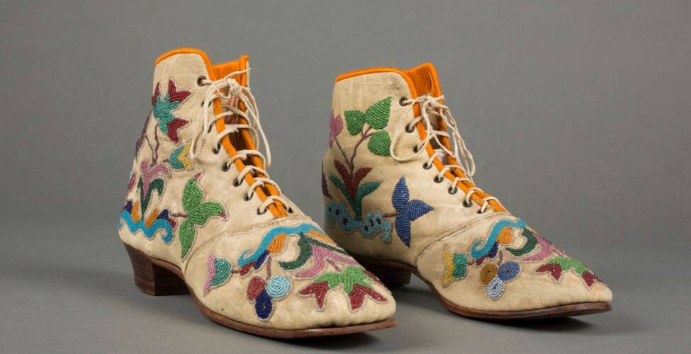 A pair of colorfully beaded, low-heeled ankle-high boot style shoes, pointing towards the right, seen in a three-quarter view against a gray background. Beige leather with beaded floral decoration on the toe box and both inner and outer sides of the shoes depicting leaves, flowers and abstract shapes in shades of blues, dark and light greens, burgundy, lilac and yellow almost cover the shoes. Beige laces thread through metal eyelets at front, the bright yellow lining is visible the length of the throat of the shoes. A seam is across the instep, they have brown soles and wooden, stacked heel. The toes of the shoes lift slightly from the smooth surface on which they are resting.