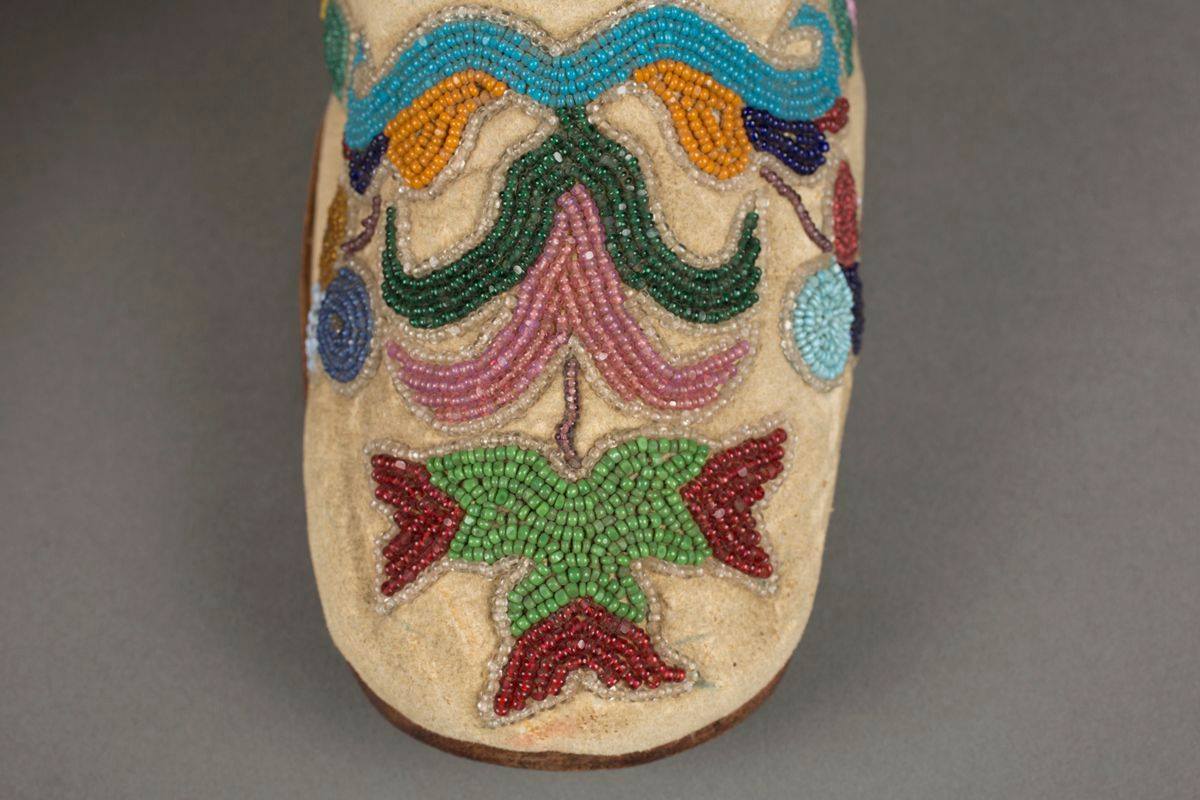 A pair of colorfully beaded, low-heeled ankle-high boot style shoes, pointing towards the right, seen in a three-quarter view against a gray background. Beige leather with beaded floral decoration on the toe box and both inner and outer sides of the shoes depicting leaves, flowers and abstract shapes in shades of blues, dark and light greens, burgundy, lilac and yellow almost cover the shoes. Beige laces thread through metal eyelets at front, the bright yellow lining is visible the length of the throat of the shoes. A seam is across the instep, they have brown soles and wooden, stacked heel. The toes of the shoes lift slightly from the smooth surface on which they are resting.