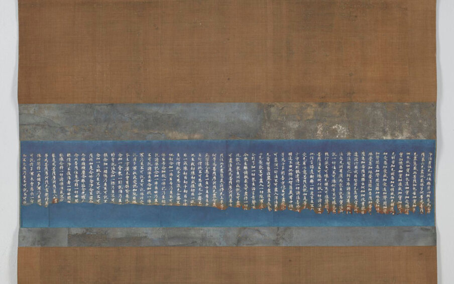 A photograph of a rectangular scroll composed of horizontal bands. Brown bands, top and bottom, appear to be of woven cloth, the top band composing about a third of the overall work. The bottom band is half the top’s width. At center is the indigo band with 56 rows of silver inked calligraphy sandwiched between the mottled metallic foil bands. At the very top are two loops for hanging and at bottom a dowel can be seen at the scroll’s edge.