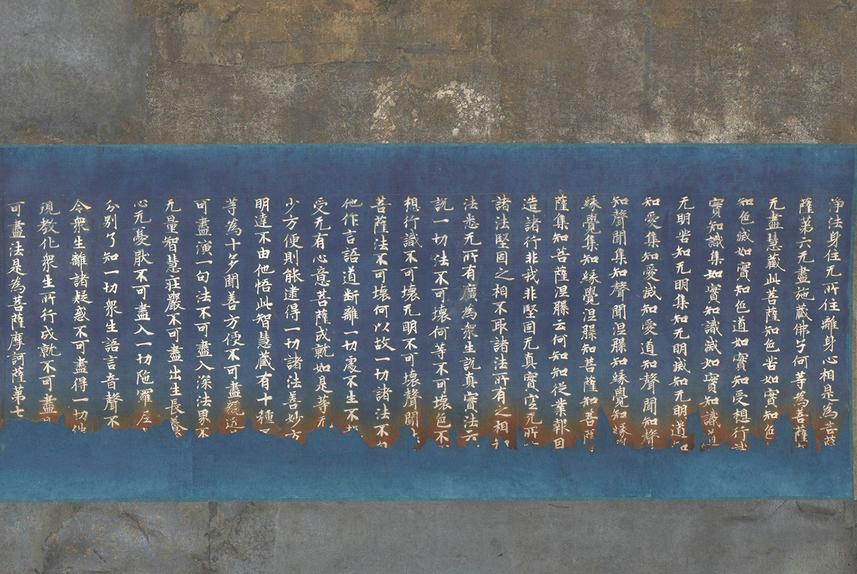 Rectangular detail of the central section of the scroll showing a wide, deep blue or indigo band running from side to side containing 28 rows of calligraphy characters in silver ink. The inked characters line up uniformly along the top of the rows but at the bottom the characters become jagged and uneven and the deep blue turns to a rusty brown. Two bands of mottled bluish-gray, taupe and cream metallic foil run horizontally along the top and bottom of the calligraphy.