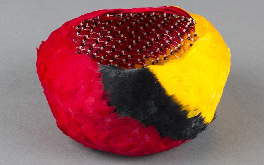 ‘ahu ‘ula, Bernice Akamine, size 2 1/2x 5 inches in diameter, dyed goose feathers, copper wire, and glass seed beads. A small, feather-clad, bowl-shaped basket shown from slightly above to provide a view inside. The outside of the basket is covered in overlapping feathers that create dense swaths of bright colors. From left to right, the colors are red, black, and yellow. The colors appear to swirl around the basket on a diagonal with each color of feather blending slightly into the next. Inside, the basket’s structure is revealed to be composed of a series of looped copper wires adorned with alternating small black and white beads. The feathers are clearly visible though the loose weave of the basket’s structure.