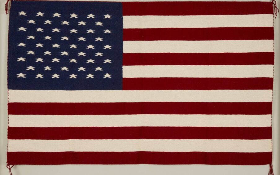 Flag rug, Bertha Harvey, 30 x 60 inches, wool. A woven American flag in traditional colors of bright red, white, and dark blue. The flag has thirteen alternating red and white horizontal stripes with a rectangle of dark blue in the upper-left corner containing fifty stars in rows. The edges are whip stitched with red yarn and the corners end in four strands of knotted red yarn.