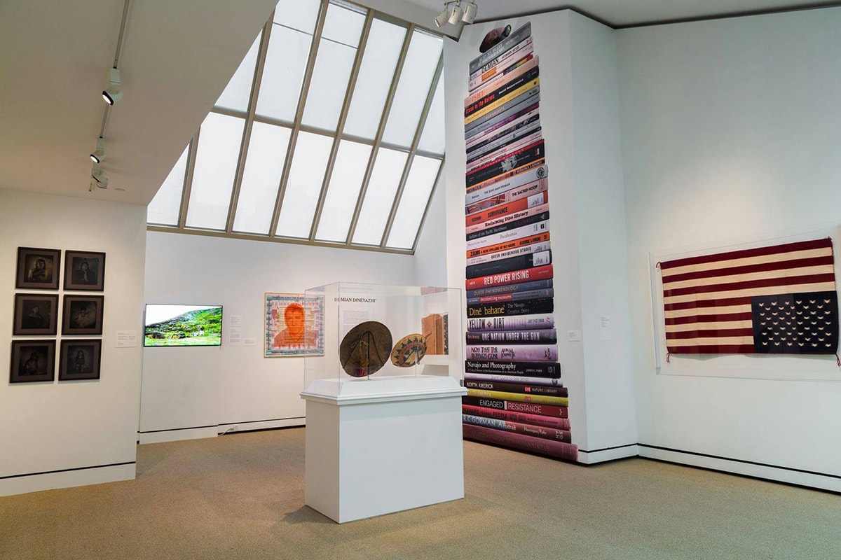 A photograph of a gallery interior featuring works of art and a large skylight window. The skylight window is at center top and extends halfway down the photo. The room has angled ceilings and walls that jut out into the room. On the wall space to the right of the skylight is an image of a stack of books that reaches from floor to the top of the double height ceiling. Some titles are: Bury My Heart at Wounded Knee, Red Power Rising. The Sacred Hoop, One Nation Under the Sun, Survivance and Yellow Dirt. To the far right and hanging on the wall is Bertha Harvey’s “Flag Rug.’ This woven, wool depiction of the American flag is hung upside down with the stars at bottom. In the center of the room is a free-standing pedestal with an acrylic enclosure containing a Native American basket and another artwork shown from the back. Behind the pedestal and below the skylight there is a video monitor showing a green landscape and a square portrait surrounded by text. The artist’s name, Demian DinéYazhi', is on the wall above a large wall label. At far left, six black-and-white portraits hang in three rows of two each. The gallery walls are white and the carpet is beige.