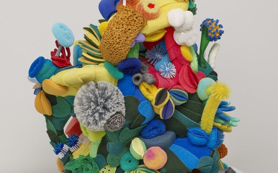 Image description: Biophilia, Lynn Aldrich, size 42 x 30 x 28 inches, sponges, brushes, scrubbers, scouring pads, mop heads, plungers, plastic, plumbing parts, wood. A sculpture composed of cleaning implements in yellows, blues, and greens with splashes of red, pink, copper, and silver and arranged to resemble a squat, asymmetrical column of sea life. Beginning at the bottom, flat dark green, blue, and yellow scrubbing pads layer to form a rounded base. Thirteen steel wool circles are applied at center forming a structure that suggests sea anemones. Blue scrubbers and dish sponges layer along with copper mesh scrubbers, nylon scrubbers in pink and green. Half circles of sponges in yellow, and shades of green are stacked at left to resemble striped plant life. A large natural light brown sea sponge features at right near red mesh scrubbers and a pink suction cup disk. Just below this, blue, green, and yellow rubber gloves are grouped at right with the fingers jutting out from sculpture. Moving up the sculpture, yellow, blue, green, and red dish sponges and pot scrubbers are folded and clustered to suggest more plant life. Plastic handled pot scrubbers protrude randomly from the sculpture. Near top left is another natural sponge next to a blue and white bottle brush. The sculpture is topped with three more bottle brushes, two white with green sponge tops and a royal blue one at left and an additional yellow dish scrubber with lavender bristles. The sculpture gives the overall sense that we could be viewing an organic life form instead of plastic cleaning implements.