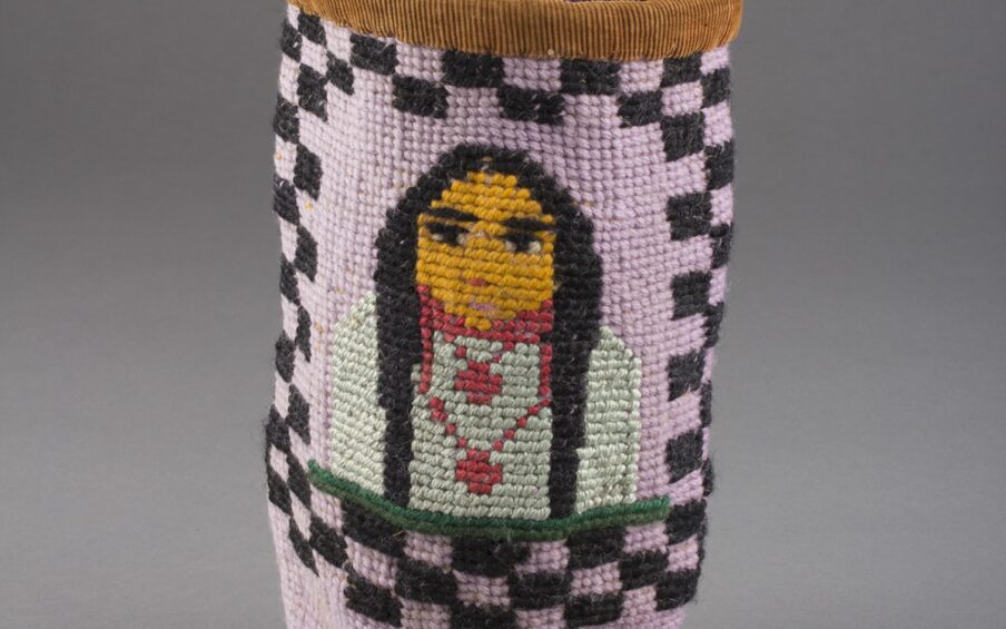 Berry bag, Umatilla artist, commercial dyes, buckskin and cotton, size: 10 x 5 ¼ inches in diameter. A tall, cylindrical, woven basket with the image of a portrait surrounded by a black checkerboard pattern. The figure is positioned at the center of the basket, showing the head and upper body. The individual has long black hair, black eyes and brows, and a golden yellow face. Two rose-colored necklaces adorn an off-white shirt. A dark green bar runs along the bottom of the figure. The body of the basket is lilac and the black checkerboard pattern surrounds the figure with two rows of check at top and sides and three rows at bottom. The rim of the basket is covered in a brown corduroy fabric. Two buckskin ties are seen knotted at the back of the rim.