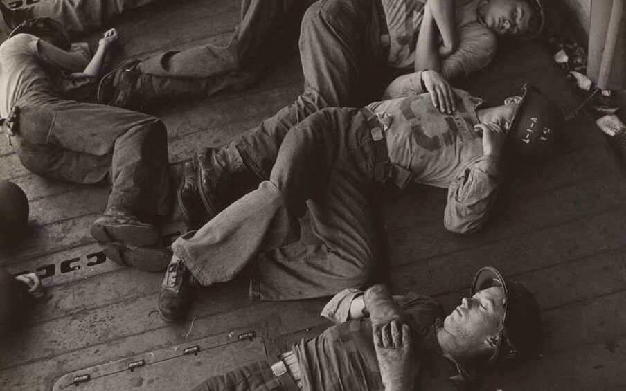 Victor Jorgenson, Untitled (relieved of their long ordeal and still in battle helmets, the exhausted crew sleeps) size: 10.5x10.5 inches, gelatin silver print. A square, black-and-white photograph of six young male soldiers lying asleep on the deck of a ship. The soldiers are viewed from above and are positioned diagonally across the photo with their heads towards the top right and feet towards the bottom left. At far left, one soldier lies on his side near the other soldier’s feet. He is positioned vertically in the photo with his head near top left. They lay on their sides with their knees drawn up or on their backs and are fully clothed in khakis, boots, and sometimes helmets. The decking beneath them runs diagonally and parallel to the men. The photo captures portions of other soldiers also sleeping suggesting a crowded deck of sleeping men.