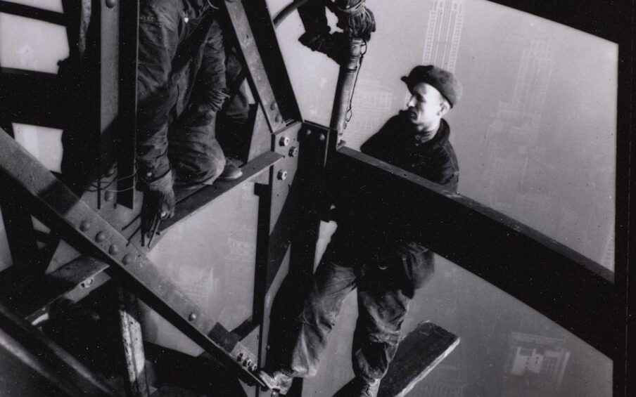 Lewis W. Hine, Top of the Mooring Mast, Empire State Building, 4 7/8 × 3 ¾ inches, gelatin silver print. A vertical rectangular, black and white photo of three light skinned men working on steel girders above a hazy cityscape. Three wide girders run parallel and diagonally from the upper left to the lower right. Other beams form a triangular structure near the upper center. One man kneels in this triangle while watching another to the right as he leans over appearing to rivet a beam below. The third man is standing at center right on a wooden plank that balances on a lower beam and juts out into the open air. The men wear work clothes of coveralls, boots, and flat caps. They do not wear safety gear. City buildings can be just made out through the haze behind them.