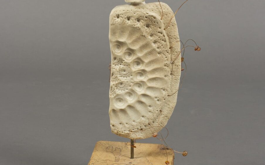Dancing in the Wind, Lillian Pitt, 14 ¾ x 5 7/8 x 4 7/8 inches, clay, beads, wood, and wire. A photo of a sculpture composed of three main parts: a body, a base, and a finial. The pale-beige clay body is shaped like a vertical rectangle with rounded corners and is highly textured with imprinted patterns. The bottom right corner is rounded off further than the other corners. The imprinted patterns appear to have been made with shells and form concentric semicircles similar to how the bands of a rainbow are arranged. A row each of scallop and nautilus shell imprints form arcs on their sides starting at the top and ending at the bottom. The balance of the body is dotted with the indentations of shell markings overall. Protruding irregularly from the right side of the body are bent copper wires with amber colored beads at the ends. Each wire is bent differently. The body is attached to a thick, chunky wood base by a short, metal rod. The blond wood of the base is traversed with thin dark lines that seem to outline the grain. At top, the finial is connected to the body by clay and is an irregular spherical shape with metal rods radiating out at 9, 12, 2, and 3 o’clock. At 10 o’clock, a copper wire curves into a spiral.
