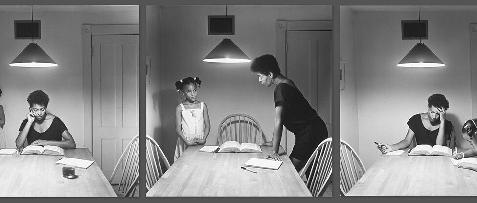 Untitled (Woman with daughter) from the series Kitchen Table, Carrie Mae Weems, each image 27 ¼ x 27 ¼ inches, gelatin silver prints. A series of three black-and-white photographs, positioned side by side, featuring a young Black girl and a Black woman in a room at a large table. The first photo shows the woman with short, dark hair and a dark dress seated at the end of a long, wooden table under a triangular overhead light. She looks down at an open book with her hand at left resting on her cheek and her arm at right resting on the thick book. Beside her to the left sit a notepad and pens. On the right side of the table is a notebook, pen, and a glass of dark liquid. To the left and behind the woman, stands a young girl in a light dress with shoulder straps, her hair up in pigtails. She stands with her arms folded and looks towards the book on the table. At right is a closed door and there are two chairs on either side of the table. The second photo shows the same setting with the woman now standing on the right side of the table leaning forward with her hands on the table. She looks directly at the girl now standing at left with hands clasped and resting on the table. The two stare intensely at each other. The third photo shows the woman back in her seat at the end of the table, left hand on her forehead, a pen in her other hand while she looks down at her book. The child is now seated at the right side of the table and is drawing in the notebook.