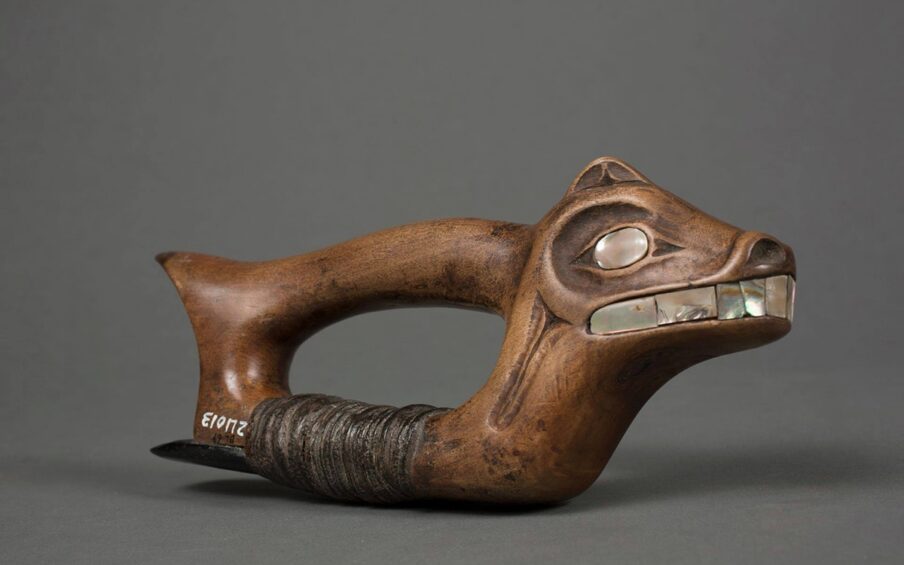 Adze, Haida artist, 4 x 9 ½ x 1 5/8 inches, wood and steatite with abalone shell inlay. A photo of a wooden adze with the head of an animal containing abalone shell facial features. The animal head is at the right side of the woodworking tool and has a small upright ear, large shell eyes, flared nostrils, and a wide mouth showing large shell teeth. The wood is a warm brown and very smooth all over. The abalone shell is a grayish silver with flashes of bright white and deeper tones of gray and brown. Extending from the back of the head to the left is the tool’s cylindrical handle that ends in a point like a short tail. The animal’s neck extends down to the base of the tool with the handle and base connecting at each end and forming a void and room for the artisan’s hand. The base is wrapped with thin strips of dark brown leather that have compacted with use. These leather strips hold a piece of a steatite to the base. The steatite extends back past the end of the adze. Old museum accession numbers are seen at the bottom rear of the base having been added years later.