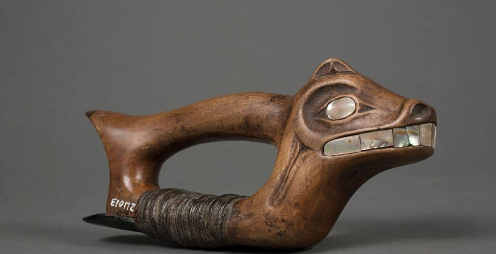 Adze, Haida artist, 4 x 9 ½ x 1 5/8 inches, wood and steatite with abalone shell inlay. A photo of a wooden adze with the head of an animal containing abalone shell facial features. The animal head is at the right side of the woodworking tool and has a small upright ear, large shell eyes, flared nostrils, and a wide mouth showing large shell teeth. The wood is a warm brown and very smooth all over. The abalone shell is a grayish silver with flashes of bright white and deeper tones of gray and brown. Extending from the back of the head to the left is the tool’s cylindrical handle that ends in a point like a short tail. The animal’s neck extends down to the base of the tool with the handle and base connecting at each end and forming a void and room for the artisan’s hand. The base is wrapped with thin strips of dark brown leather that have compacted with use. These leather strips hold a piece of a steatite to the base. The steatite extends back past the end of the adze. Old museum accession numbers are seen at the bottom rear of the base having been added years later.