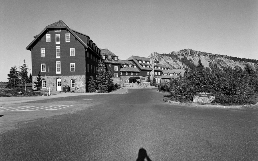 Crater Lake Lodge, Jonathan Calm, pigment print. A horizontal rectangular, black-and-white photo of the Crater Lake Lodge buildings and its driveway entrance. The top half of the photo features the lodge seen from one end and running diagonally back and across the photo’s middle ground. The buildings that compose the lodge are faced in light-colored stonework from the ground to just above door height with dark cladding finishing the rest of the structures. Rows of white-paned windows dot the attached buildings that are of varying heights. To the left of the lodge is a rocky hilltop with sparse vegetation and to the right are several evergreen trees. The bottom half of the photo shows the expansive blacktop driveway that extends out from the lodge entrance and to each side. At bottom center is the photographer's shadow, shown from chest up. At center far right, shrubbery backs a wooden sign mounted on a stone platform. The sign reads: ”Crater Lake Lodge”.