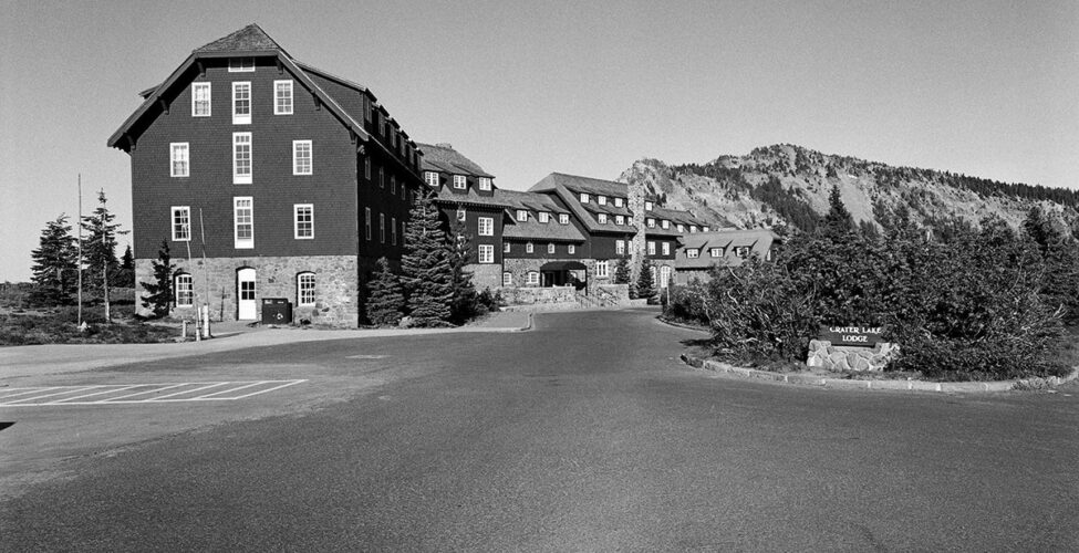 Crater Lake Lodge, Jonathan Calm, pigment print. A horizontal rectangular, black-and-white photo of the Crater Lake Lodge buildings and its driveway entrance. The top half of the photo features the lodge seen from one end and running diagonally back and across the photo’s middle ground. The buildings that compose the lodge are faced in light-colored stonework from the ground to just above door height with dark cladding finishing the rest of the structures. Rows of white-paned windows dot the attached buildings that are of varying heights. To the left of the lodge is a rocky hilltop with sparse vegetation and to the right are several evergreen trees. The bottom half of the photo shows the expansive blacktop driveway that extends out from the lodge entrance and to each side. At bottom center is the photographer's shadow, shown from chest up. At center far right, shrubbery backs a wooden sign mounted on a stone platform. The sign reads: ”Crater Lake Lodge”.