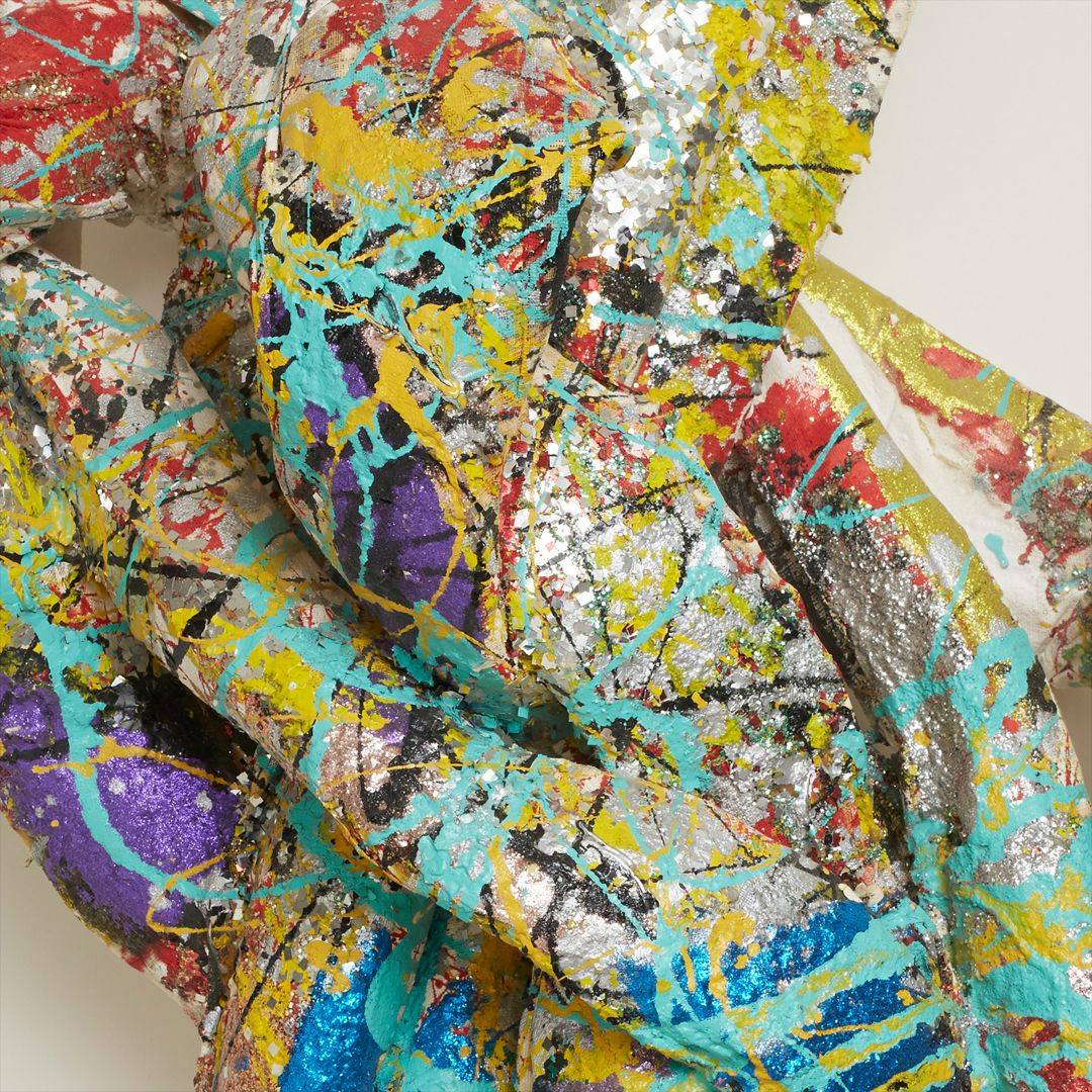 A closeup photo of the sculpture showing the structure of the knot and splatter work. Bright colors and glitter cover the thick strands that compose the sculpture.
