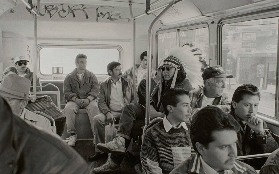 Indian Man on the Bus, from the series Indian Man in San Francisco, 16 x 20 inches, gelatin silver print. A horizontal, rectangular, black and white photo of the crowded interior of a bus featuring a man wearing a Native American feather headdress. The view is of the last few rows of the back of the bus showing a group of seated male presenting individuals. At right, a man is seen wearing a large Native American feather headdress composed of dark tipped, white feathers that radiate from a beaded band across the top of the man’s forehead. He wears a neutral expression, dark sunglasses, a jacket, cuffed jeans and high-top sneakers. He is sitting on one of the inward facing bus benches and so faces towards the left. The other figures around him sit in rows: three behind the man in the headdress, across the back wall of the bus, four seated in rows in front of the man and several partially visible figures to the left of the photo. The men, all of whom look past the man in the headdress or out the window, appear to be of varying backgrounds such as White, Hispanic or Asian. A city street scene is visible through large bus windows at right.
