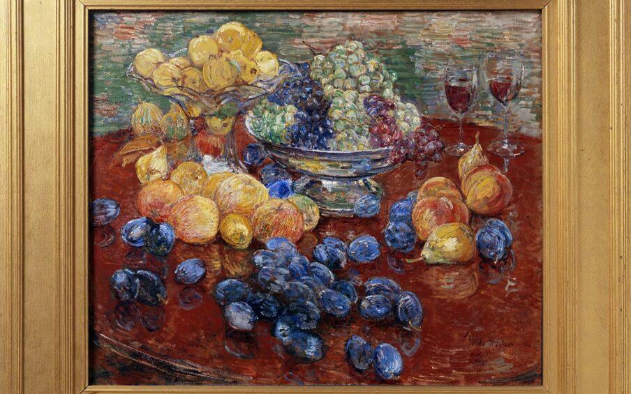 Childe Hassam. Oregon Still Life, 25 x 30 ¼ inches, oil on canvas. A painting of an assortment of fruit laid out on a round, dark brown table, the edges of which are mostly out of frame. At top, at 11 and 12 o’clock, are a glass bowl with a tall stand brimming with yellow apples that sits beside a squat, silver bowl holding green, purple, and red grapes. Just below the glass bowl are more yellow apples, some with a hint of pink. Further down the painting and scattered over the table are deep blueish-purple plums. At three o’clock are a grouping of a half dozen peaches and pears set near two half-filled glasses that suggest wine. The painting technique is bold and uses dry brush strokes. Paint is heavily layered, revealing the colors underneath in areas. The table and fruit are set against a background heavily painted with horizontal strokes in muted greens, terra cotta, and whites with deep yellow.