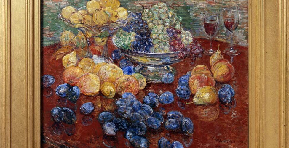 Childe Hassam. Oregon Still Life, 25 x 30 ¼ inches, oil on canvas. A painting of an assortment of fruit laid out on a round, dark brown table, the edges of which are mostly out of frame. At top, at 11 and 12 o’clock, are a glass bowl with a tall stand brimming with yellow apples that sits beside a squat, silver bowl holding green, purple, and red grapes. Just below the glass bowl are more yellow apples, some with a hint of pink. Further down the painting and scattered over the table are deep blueish-purple plums. At three o’clock are a grouping of a half dozen peaches and pears set near two half-filled glasses that suggest wine. The painting technique is bold and uses dry brush strokes. Paint is heavily layered, revealing the colors underneath in areas. The table and fruit are set against a background heavily painted with horizontal strokes in muted greens, terra cotta, and whites with deep yellow.