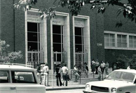 Horizontal, rectangular black-and-white photo of the front of the Museum’s entrance, three vertical, evenly spaced entryways faced in light marble with 3 windows each above. 15 people are gathered on the steps, standing and sitting at ease. In the foreground 2 cars are parked along the street.