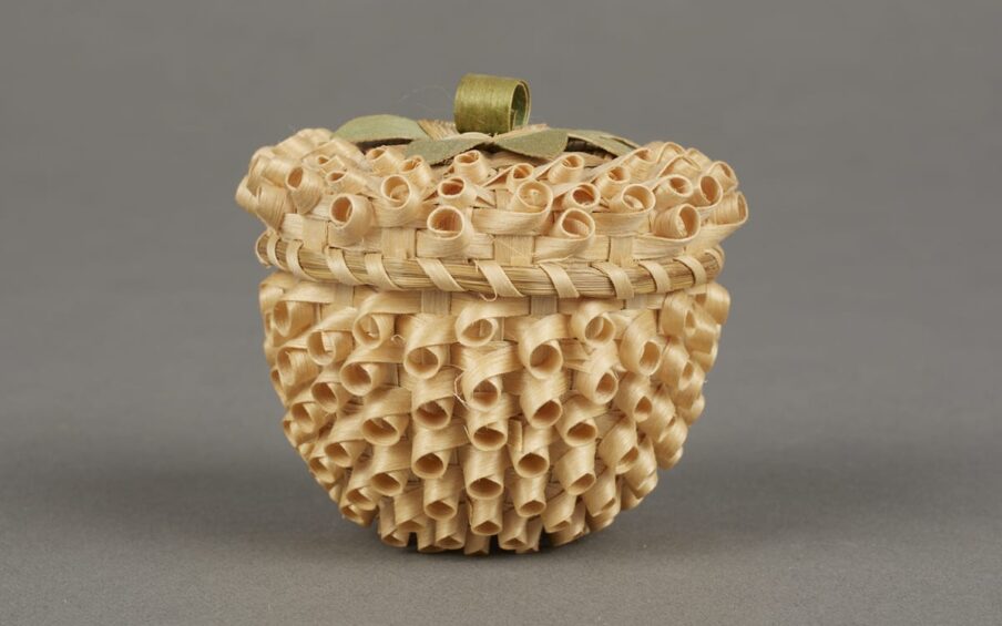 Strawberry Basket by Mary Adams (Mohawk, Canadian), 2 inches x 2 inches, ash splints and commercial die. This tiny basket with a lid is a natural golden color overall and topped with a decorative loop handle that is dyed green to imitate leaves and a stem. The whole basket has a bumpy texture due to the weave of the materials. The weave creates rows of small looping circles that hug the surface with the open ends of the loops facing outward. They are tilted slightly downward on the body of the basket and more upward on the lid. Around the edge of the lid, where it meets the body of the basket, the weaving materials are bound together with a splint spiralling around the edge. It looks very much like an unripe strawberry with the dark shadowy centers of each of the loops resembling rows of seeds.