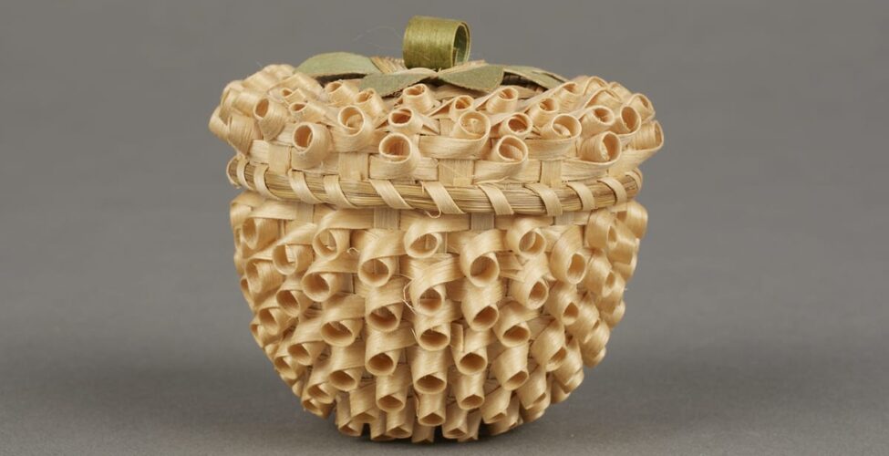 Strawberry Basket by Mary Adams (Mohawk, Canadian), 2 inches x 2 inches, ash splints and commercial die. This tiny basket with a lid is a natural golden color overall and topped with a decorative loop handle that is dyed green to imitate leaves and a stem. The whole basket has a bumpy texture due to the weave of the materials. The weave creates rows of small looping circles that hug the surface with the open ends of the loops facing outward. They are tilted slightly downward on the body of the basket and more upward on the lid. Around the edge of the lid, where it meets the body of the basket, the weaving materials are bound together with a splint spiralling around the edge. It looks very much like an unripe strawberry with the dark shadowy centers of each of the loops resembling rows of seeds.