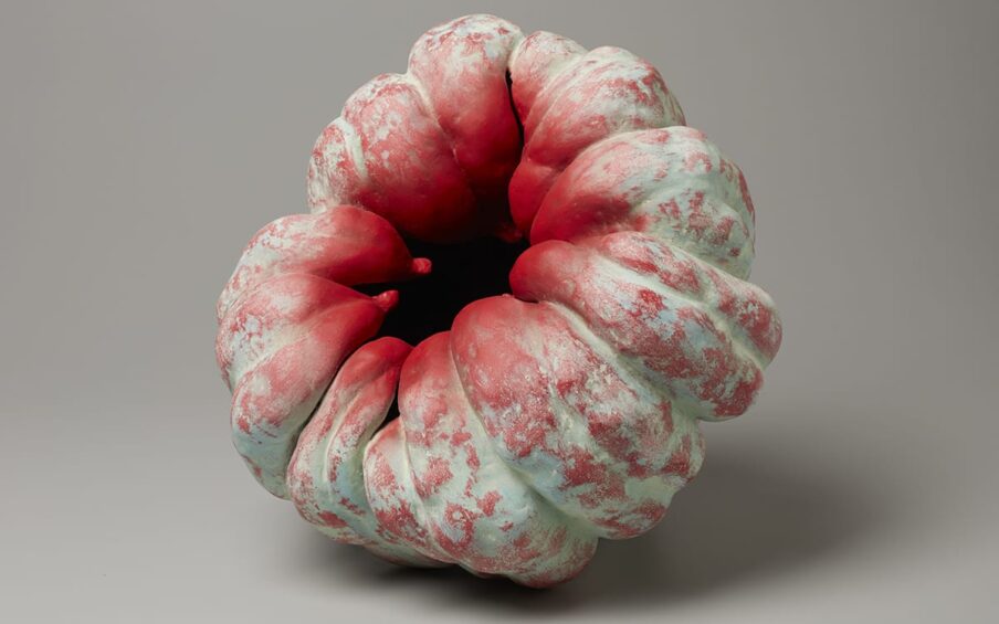Akoda (Pumpkin), Katsumata Chieko, 16 ⅛ inches x 16 ⅛ inches x 16 ⅛ inches, stoneware with pigment. The ceramic object is pumpkin-like, laying on its side at a ¾ turn to the left. It has bulbous, ridged sections that wrap around to the center, which is open and caving in on itself. The bulging sections seem to taper off to small nubs and become freed from each other as they curl inward towards the dark hollow at the center. There is a pale, minty green underlayer of pigment with bright red pigment applied here and there to the bumpy ridges that gradually increases until all around the opening is completely red.
