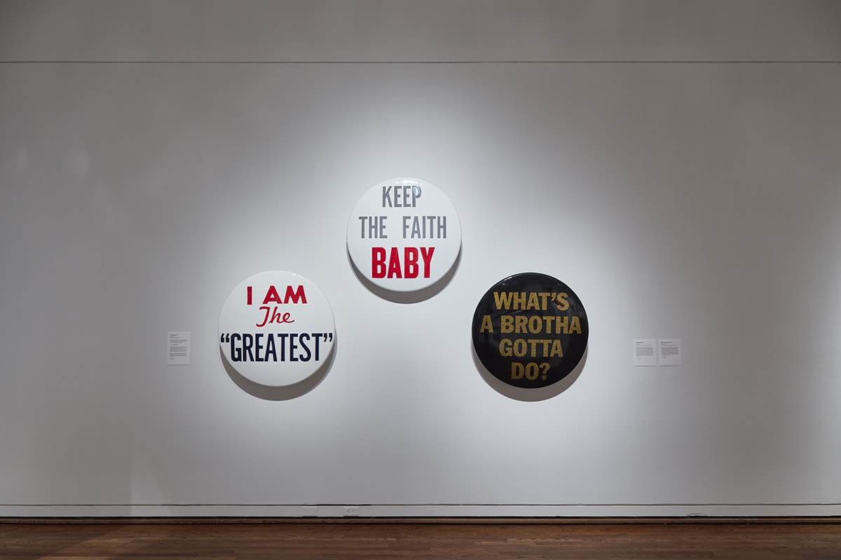 Keep the Faith, Hank Willis Thomas, each 34 inches in diameter, fiberglass and aluminum. A photo of three oversized round metal objects meant to look like political buttons with slogans hanging on a gallery wall. Each button has a different slogan, colorways and fonts. Two are hung at eye level with the third in the middle hung slightly higher. The first at left reads: I am the greatest” on a white background. “I am” is capitalized in red font. “The” is in italicized in red with a capital T. “The greatest” is capitalized in navy blue and has quotation marks. The middle button reads ”Keep the faith baby” on a white background. All words are capitalized with “baby” in bold red font and the rest in pale grey font. The last button at right reads “What’s a brotha gotta do?” in gold, capitalized font on a black background. The three objects hang on a white wall with gallery labels at far left and far right of the art. A narrow strip of wooden floor is visible at the lower edge.