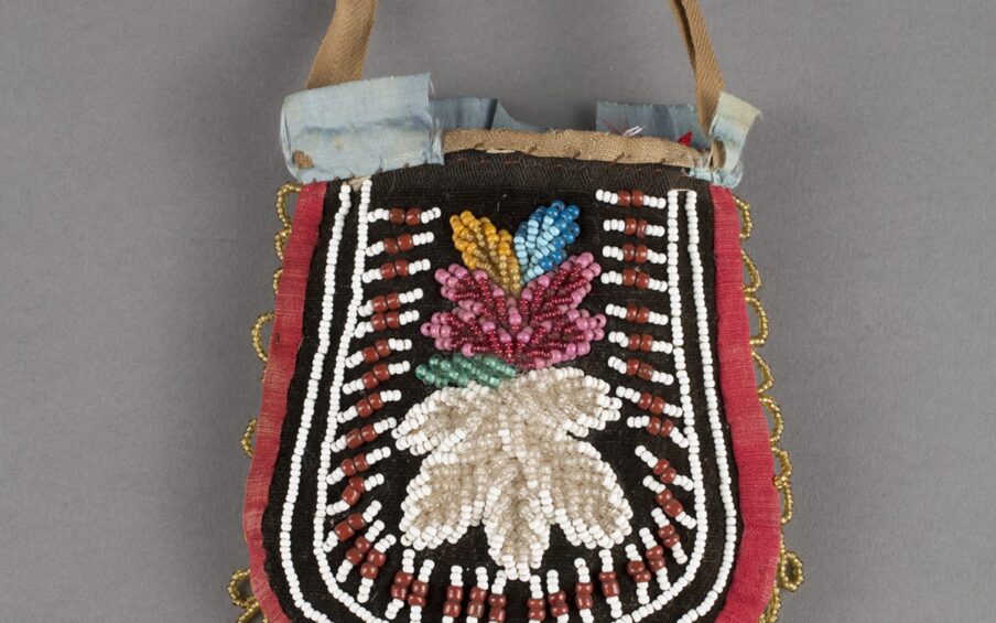 Haudenosaunee/Iroquois artist, Pouch, 8 x 4 inches, cotton, silk and glass beads. A photo of a colorful U-shaped pouch with handle against a gray background. The pouch is slightly narrower at the top then widens at the bottom. At the center, set against black velvet is a large, white, beaded flower topped by a pink, beaded flower with pale green, gold, and blue, beaded leaves. Surrounding the flowers are white and brick-red beadwork lines composed of three white beads, two larger brick-red beads, then three white beads. Each line radiates outward from the flowers at center. Two lines of white beading encompasses the center designs running along the edge of the pouch. At the outer edge of the pouch is a red cotton border with loops of gold beads along the sides and turquoise blue beaded loops along the bottom. Some areas of beading are missing at the bottom center and at the left edge. The top of the pouch shows a thin, short, tan cotton handle. Pale blue ribbon embellishes the opening of the pouch but is partially missing, exposing the hand stitching along the pouch’s opening.