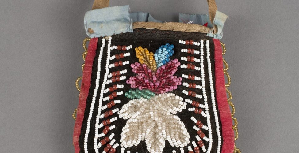 Haudenosaunee/Iroquois artist, Pouch, 8 x 4 inches, cotton, silk and glass beads. A photo of a colorful U-shaped pouch with handle against a gray background. The pouch is slightly narrower at the top then widens at the bottom. At the center, set against black velvet is a large, white, beaded flower topped by a pink, beaded flower with pale green, gold, and blue, beaded leaves. Surrounding the flowers are white and brick-red beadwork lines composed of three white beads, two larger brick-red beads, then three white beads. Each line radiates outward from the flowers at center. Two lines of white beading encompasses the center designs running along the edge of the pouch. At the outer edge of the pouch is a red cotton border with loops of gold beads along the sides and turquoise blue beaded loops along the bottom. Some areas of beading are missing at the bottom center and at the left edge. The top of the pouch shows a thin, short, tan cotton handle. Pale blue ribbon embellishes the opening of the pouch but is partially missing, exposing the hand stitching along the pouch’s opening.