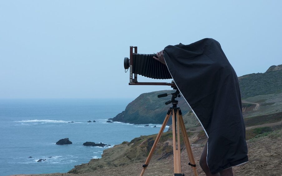 Double Vision, Jonathan Calm, pigment print. A vertical rectangular photograph of man using a camera on a tripod near an oceanside cliff. The man is seen from the side, his camera pointed to the left. The camera has a large black bellows attached to a lens and stands on light colored wooden tripod legs. The man is mostly concealed under a dark cloth with only his bare legs and the fingers of one hand visible. He is dark skinned and barefoot. He stands on rough, brown ground with cliffs and the ocean behind his. The upper half of the photo contains a pale blue sky.