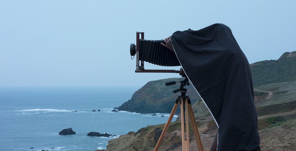 Double Vision, Jonathan Calm, pigment print. A vertical rectangular photograph of man using a camera on a tripod near an oceanside cliff. The man is seen from the side, his camera pointed to the left. The camera has a large black bellows attached to a lens and stands on light colored wooden tripod legs. The man is mostly concealed under a dark cloth with only his bare legs and the fingers of one hand visible. He is dark skinned and barefoot. He stands on rough, brown ground with cliffs and the ocean behind his. The upper half of the photo contains a pale blue sky.