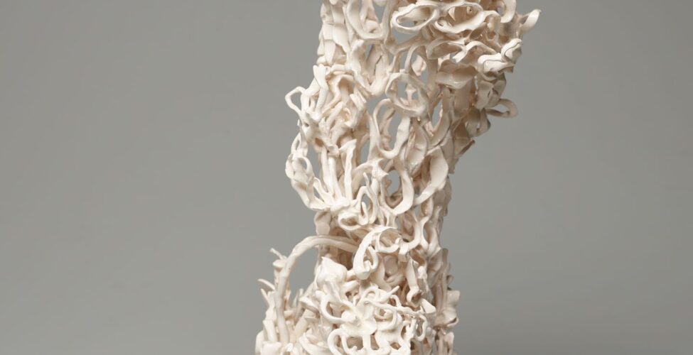 An open-work, vertical sculpture evoking the shape of a vase in creamy white. The surface is composed of oval clay loops that connect irregularly to create a highly textured lace-like body that sits on four folded clay coils. The structure appears to lean slightly to the left in this view and shows clay loops both crowded and compressed in areas like upper right and stretched and spaced in the lower left part of the sculpture. This gives the work an organic quality of perhaps coral or a sea creature.