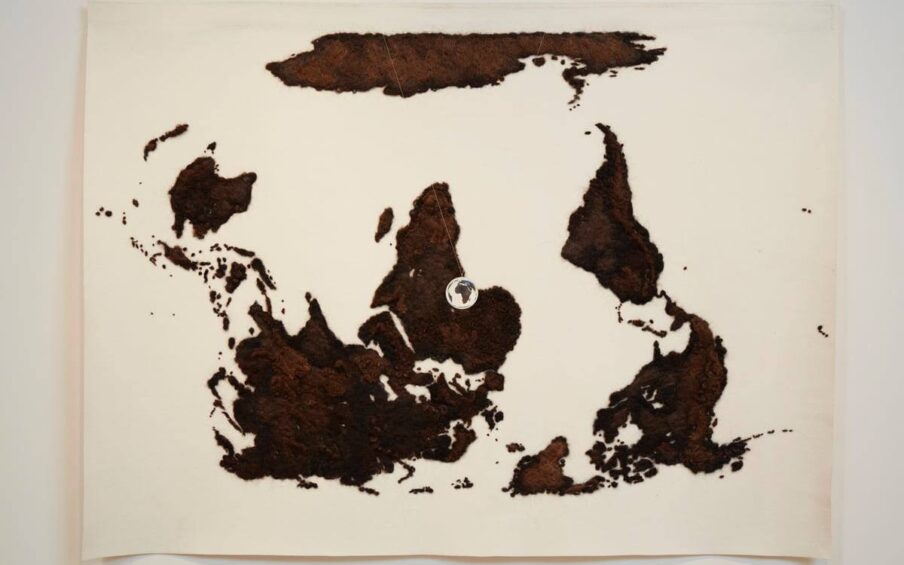 A landscape-oriented work of a flat map of the world rendered in brown human hair on a cream-colored wool felt ground. The map is upside down so Antarctica is across the top, North and South America are at right, Australia is at left and Africa is in the center. The land masses are rendered in brown hues of human hair giving them a fuzzy, textured appearance. Viewing the map upside down makes it appear to be an abstract work of scattered large and small irregular shapes. Suspended in front of the map is a small, clear acrylic ball through which the continent of Africa can be seen, right side up.