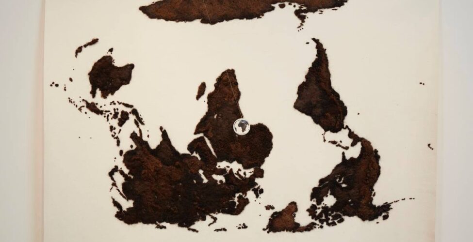 A landscape-oriented work of a flat map of the world rendered in brown human hair on a cream-colored wool felt ground. The map is upside down so Antarctica is across the top, North and South America are at right, Australia is at left and Africa is in the center. The land masses are rendered in brown hues of human hair giving them a fuzzy, textured appearance. Viewing the map upside down makes it appear to be an abstract work of scattered large and small irregular shapes. Suspended in front of the map is a small, clear acrylic ball through which the continent of Africa can be seen, right side up.