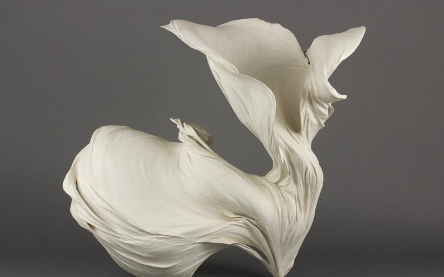 Flow #1. Fujikasa Satoko, 2011. Stoneware with matte white slip. 26 ⅛ inches high x 27 inches wide x 23 ⅛ inches deep. This sculpture resembles a light, flowing fabric garment billowing weightlessly in the wind. The sculpture touches the display surface in two points. The contact on the right is small and pointed. The material appears to flow upward and outward to the left from this point. The material flowing to the left twists and unfolds into a large round billow that has a strong sense of movement. The material moving upward from the point flows up in a thin, vertical flow that twists and opens into a funnel shape with multiple layers unfolding along the outer edges.
